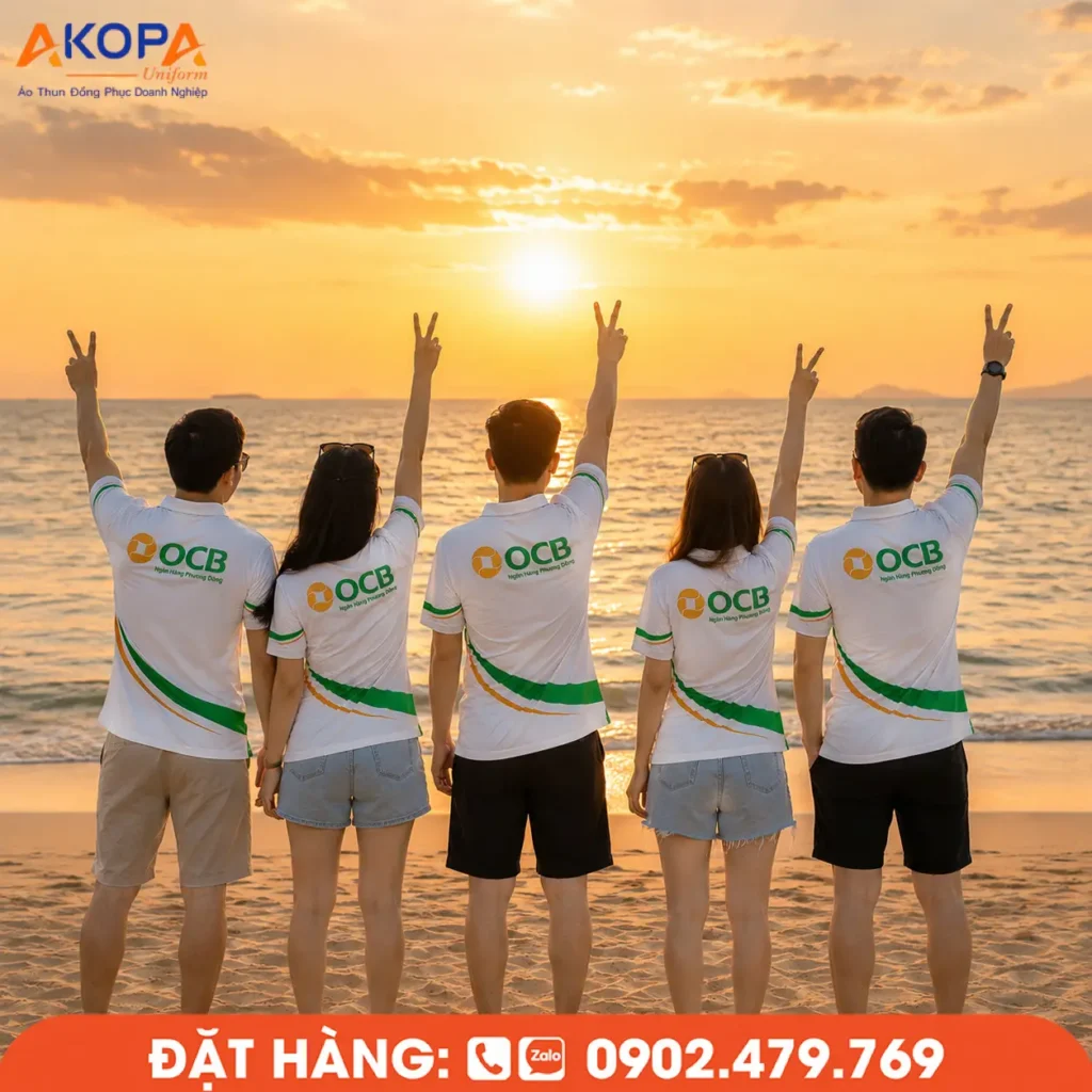 Áo polo team building màu trắng phối xanh nổi bật, thiết kế in logo sau lưng giúp tập thể dễ nhận diện khi tham gia du lịch biển. Hình ảnh cả đội cùng giơ tay trước bình minh tạo cảm giác gắn kết, trẻ trung và đầy năng lượng.