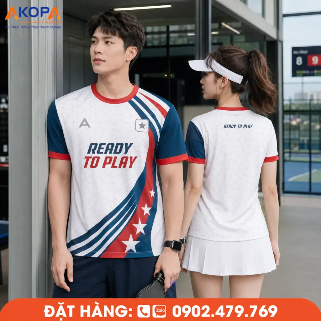 Áo đồng phục pickleball cổ tròn tông trắng phối xanh navy và đỏ nổi bật, thiết kế năng động với slogan "Ready To Play", chất liệu thoáng khí, co giãn tốt, phù hợp cho thi đấu, luyện tập và đồng phục câu lạc bộ Pickleball.